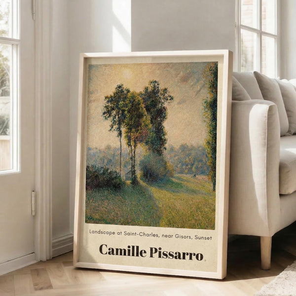May include: Framed art print of an impressionist landscape painting featuring tall trees along a winding path at sunset, displayed in a bright living room corner next to a beige armchair. The artwork shows green foliage and golden sky in soft, textured brushstrokes within a light wooden frame against white walls and hardwood floors.