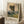 May include: Framed reproduction of an impressionist landscape painting showing tall trees along a winding country path at sunset, with soft yellows, greens, and blues. The beige-matted print leans against a white wall next to a coffee cup on wooden books and a cream blanket on a textured white rug.