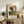 May include: Framed Impressionist landscape poster featuring tall trees in a pastoral setting with warm golden and green tones, displayed on a rustic wooden sideboard alongside potted fern, ceramic vase, and books in a cozy living room with beige chair and neutral walls.