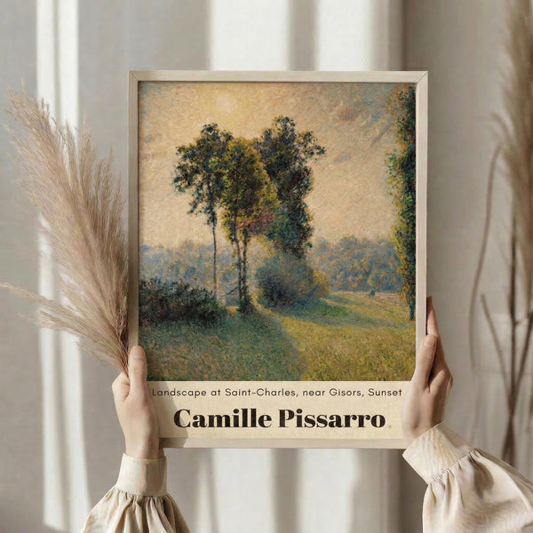 May include: Person in beige clothing holding framed art print showing impressionist landscape painting with tall trees, rolling hills, and winding path in soft greens and blues. Dried pampas grass stems visible on left side against neutral curtained background.