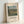 May include: Framed art print featuring a pastoral countryside landscape with rolling green fields, farm buildings, and tall cypress trees under a cloudy blue sky. The vintage-style poster displays "Landscape at Les Pâtis, Pontoise" and "Camille Pissarro" text below the impressionist artwork. Natural wood frame with cream matting, leaning against white wall corner on light wooden surface.