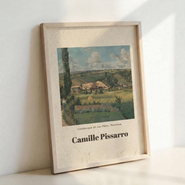 May include: Framed art print featuring a pastoral countryside landscape with rolling green fields, farm buildings, and tall cypress trees under a cloudy blue sky. The vintage-style poster displays "Landscape at Les Pâtis, Pontoise" and "Camille Pissarro" text below the impressionist artwork. Natural wood frame with cream matting, leaning against white wall corner on light wooden surface.