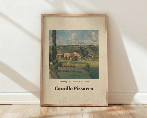May include: Framed art print leaning against white wall on light wood floor with natural sunlight casting shadows. Features vintage landscape painting showing rural countryside with rolling green fields, farm buildings with orange roofs, tall poplar tree, and cloudy blue sky. Print has cream colored mat border with artist name in bold black text below the image, displayed in thin natural wood frame.
