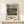 May include: Framed art print of Camille Pissarro's pastoral landscape painting featuring rolling green hills, farmhouses, and cloudy sky, displayed in light wood frame on white wooden shelf next to small wrapped gift with evergreen sprig, against cream wall