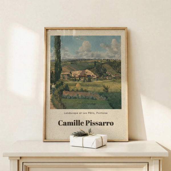 May include: Framed art print of Camille Pissarro's pastoral landscape painting featuring rolling green hills, farmhouses, and cloudy sky, displayed in light wood frame on white wooden shelf next to small wrapped gift with evergreen sprig, against cream wall