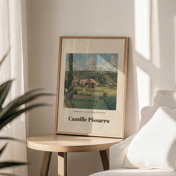 May include: Framed art print featuring a pastoral landscape painting with rolling green hills, farm buildings, and cloudy blue sky, displayed on a round wooden side table next to a beige upholstered chair. Sunlight streams across the neutral-toned room, casting soft shadows from a plant's leaves. The print shows an impressionist-style countryside scene in muted greens and earth tones within a thin wooden frame.