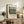 May include: Framed art print of Camille Pissarro's pastoral landscape painting showing rolling green countryside with farm buildings and cloudy blue sky, displayed on rustic wooden sideboard alongside fern plant, stacked books, ceramic vase, and brown leather chair in neutral-toned living room setting.
