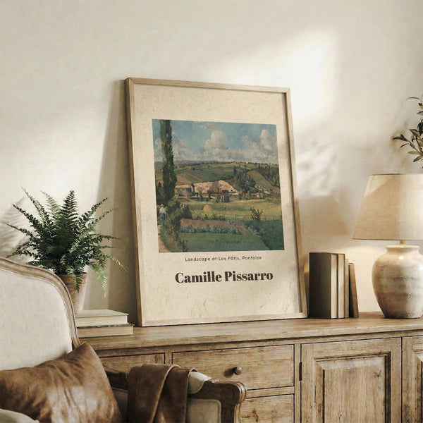 May include: Framed art print of Camille Pissarro's pastoral landscape painting showing rolling green countryside with farm buildings and cloudy blue sky, displayed on rustic wooden sideboard alongside fern plant, stacked books, ceramic vase, and brown leather chair in neutral-toned living room setting.