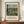 May include: A framed vintage art poster displaying a pastoral countryside landscape painting with rolling green hills, farm buildings, and cloudy blue sky. The beige poster features the artist name "Camille Pissarro" in brown text below the artwork. The brown wooden frame sits on a light wood surface beside a white ceramic vase and stacked books against cream wall paneling.