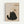 A minimalist illustration of a black cat with yellow eyes sitting upright, looking up at a ball of yarn dangling from a wooden platform above. The cat has a textured, sketchy appearance against a beige background. "Meow" is written in small text at the bottom left. The artwork is displayed in a light wood frame on a white wall.