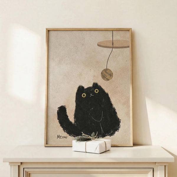 May include: Framed poster displaying a black fluffy cat with golden eyes sitting beneath a dangling yarn ball, with "Meow" text below. Artwork has textured beige background in natural wood frame, displayed on light wooden console table alongside small wrapped gift with dried botanicals against cream wall.