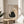 May include: Framed art print displayed on round wooden side table next to beige upholstered chair and plant. The artwork features a black fuzzy cat with yellow eyes reaching up toward a dangling ball of yarn, with "Meow" text below. Light wooden frame, neutral beige background, soft natural lighting creates shadows across the minimalist room setting.