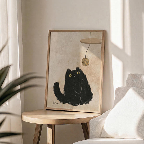 May include: Framed art print displayed on round wooden side table next to beige upholstered chair and plant. The artwork features a black fuzzy cat with yellow eyes reaching up toward a dangling ball of yarn, with "Meow" text below. Light wooden frame, neutral beige background, soft natural lighting creates shadows across the minimalist room setting.