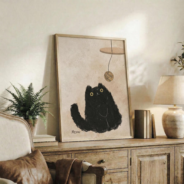May include: Framed poster print displayed on rustic wooden sideboard showing minimalist black cat illustration with yellow eyes playing with dangling yarn ball, surrounded by fern plant, ceramic vases, stacked books, and leather chair in warm neutral-toned living room setting