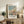 May include: Framed Monet beach scene poster with people in period dress on sandy shore, displayed on rustic wooden console table beside brown leather chair, fern plant, and ceramic vase in neutral-toned living room setting.