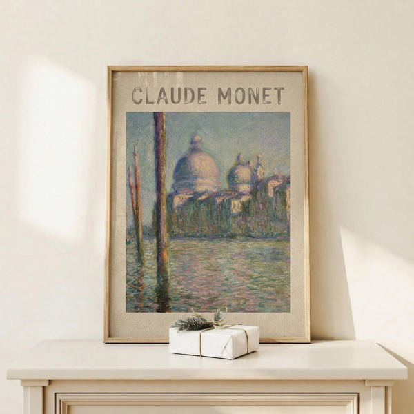 May include: Framed Monet poster print showing Venice's San Giorgio Maggiore church with distinctive domes reflected in canal water, painted in impressionist style with soft blues, greens and pinks. Displayed on light wooden surface with small white wrapped gift beside frame. Natural lighting creates gentle shadows on neutral wall background.