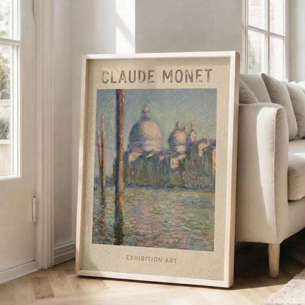 May include: Framed art print showing Monet's impressionist painting of Venice's San Giorgio Maggiore church with distinctive domes across shimmering water, with wooden posts in foreground. Print has cream matting with "CLAUDE MONET" and "EXHIBITION ART" text, displayed in light wood frame leaning against white wall next to beige upholstered chair in bright, naturally-lit room with white wooden floors.