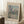 May include: Framed exhibition poster featuring Claude Monet's impressionist painting of Venice's San Giorgio Maggiore church with distinctive domes and bell towers reflected in shimmering water, rendered in soft blues, pinks and greens. The beige poster has cream matting and light wood frame, leaning against white paneled walls beside a cozy reading nook with textured throw blanket, wooden side table holding white coffee cup and saucer on white shag rug.
