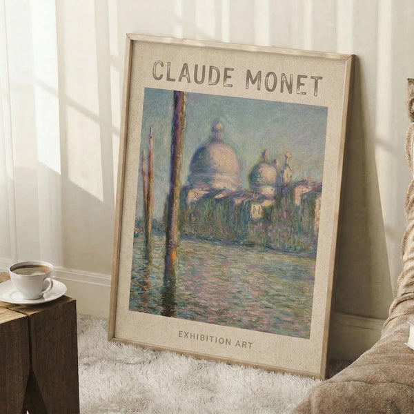 May include: Framed exhibition poster featuring Claude Monet's impressionist painting of Venice's San Giorgio Maggiore church with distinctive domes and bell towers reflected in shimmering water, rendered in soft blues, pinks and greens. The beige poster has cream matting and light wood frame, leaning against white paneled walls beside a cozy reading nook with textured throw blanket, wooden side table holding white coffee cup and saucer on white shag rug.