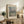 May include: Framed Claude Monet exhibition poster featuring Venice's San Giorgio Maggiore church with domed architecture reflected in water, painted in impressionist style with soft blues, greens, and warm tones. The poster sits on a rustic wooden console alongside ferns, books, ceramic vases, and throw pillows in a neutral-toned living room setting.