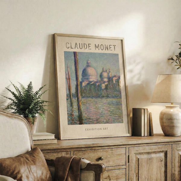 May include: Framed Claude Monet exhibition poster featuring Venice's San Giorgio Maggiore church with domed architecture reflected in water, painted in impressionist style with soft blues, greens, and warm tones. The poster sits on a rustic wooden console alongside ferns, books, ceramic vases, and throw pillows in a neutral-toned living room setting.