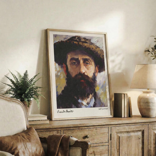 May include: Framed art print of bearded man wearing wide-brimmed hat, displayed on rustic wooden console table alongside fern plant, ceramic vase, and stacked books. Warm neutral living room setting with cream upholstered chair visible. Print features rich oil painting style with earthy tones and expressive brushwork in natural wood frame.