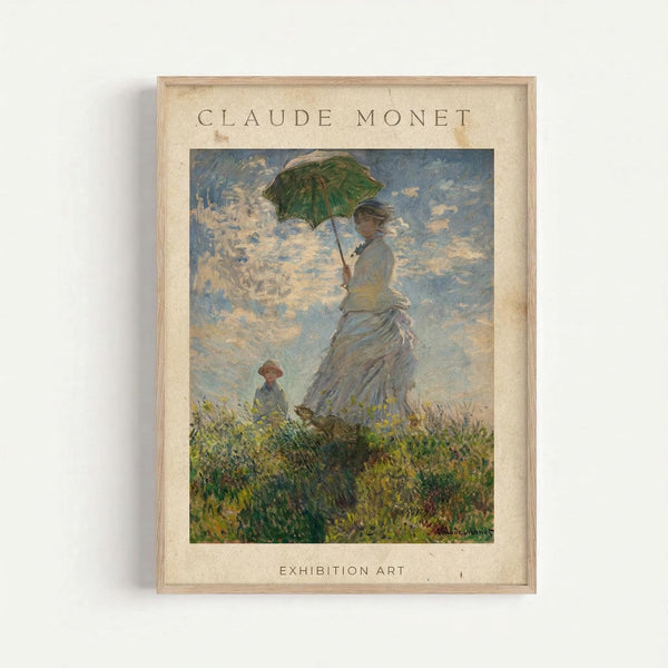 A framed reproduction of Claude Monet's impressionist painting showing a woman in a white blouse and long skirt holding a green parasol, standing on a grassy hillside with a small child nearby. The sky features soft blue and white clouds painted in Monet's characteristic loose brushstroke style. The print has "CLAUDE MONET" at the top and "EXHIBITION ART" at the bottom on cream-colored matting, displayed in a light wood frame against a white wall.