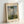 May include: Framed reproduction of Monet's impressionist painting showing a woman in a flowing white dress holding a green parasol, with a small child visible in tall grass below. The artwork features soft blues and whites in the cloudy sky with loose, painterly brushstrokes characteristic of impressionism. The print sits in a light wood frame against a white wall, casting subtle shadows.