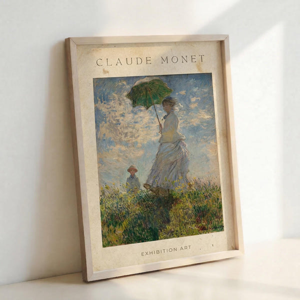 May include: Framed reproduction of Monet's impressionist painting showing a woman in a flowing white dress holding a green parasol, with a small child visible in tall grass below. The artwork features soft blues and whites in the cloudy sky with loose, painterly brushstrokes characteristic of impressionism. The print sits in a light wood frame against a white wall, casting subtle shadows.