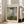 May include: Framed Claude Monet reproduction poster showing woman in white dress holding green parasol with small child in grassy field under cloudy blue sky. Cream-colored mat with "CLAUDE MONET" and "EXHIBITION ART" text. Light wooden frame leaning against white wall next to beige upholstered chair in bright room with large windows and natural light casting shadows on wooden floor.