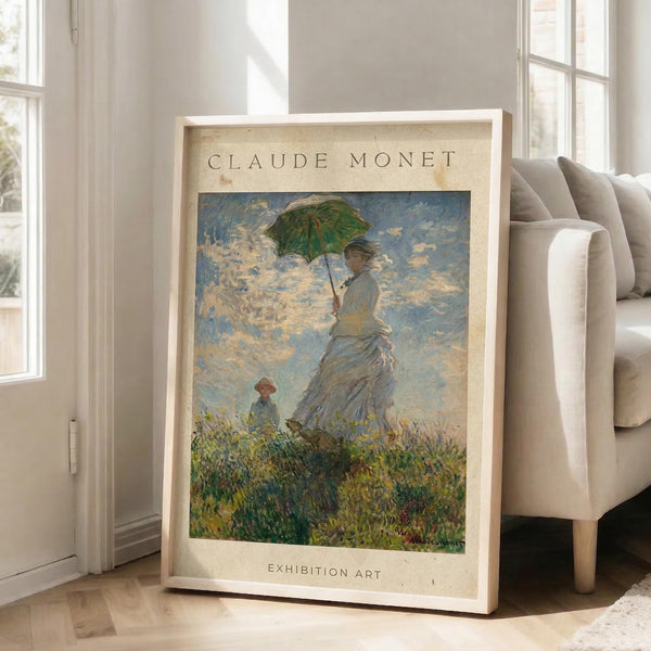 May include: Framed Claude Monet reproduction poster showing woman in white dress holding green parasol with small child in grassy field under cloudy blue sky. Cream-colored mat with "CLAUDE MONET" and "EXHIBITION ART" text. Light wooden frame leaning against white wall next to beige upholstered chair in bright room with large windows and natural light casting shadows on wooden floor.