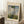 May include: Framed Claude Monet exhibition poster leaning against white wall corner on textured cream rug. Shows impressionist painting of woman in flowing white dress holding green parasol, with child figure in grassy meadow under cloudy blue sky. Warm beige mat border with vintage-style text. Coffee cup on wooden block nearby, soft natural window lighting creates shadows.
