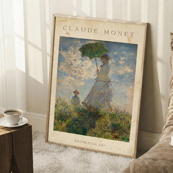 May include: Framed Claude Monet exhibition poster leaning against white wall corner on textured cream rug. Shows impressionist painting of woman in flowing white dress holding green parasol, with child figure in grassy meadow under cloudy blue sky. Warm beige mat border with vintage-style text. Coffee cup on wooden block nearby, soft natural window lighting creates shadows.