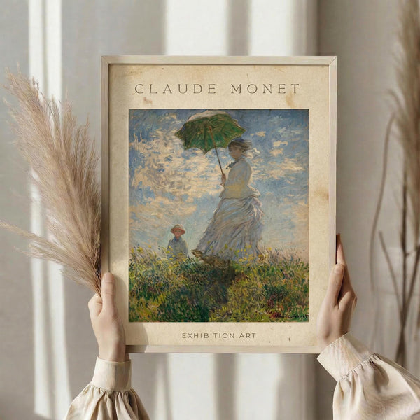 May include: Hands holding a framed vintage-style art print featuring Monet's impressionist painting of a woman in a white dress holding a green parasol, with a child in a grassy field under cloudy skies. The beige matted frame shows "CLAUDE MONET" and "EXHIBITION ART" text. One hand also holds dried wheat stems against a soft neutral background with white curtains.