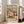 May include: Framed vintage-style beach scene poster leaning against white wall next to beige sofa in bright living room. Artwork shows crowded sandy beach with pink and blue striped umbrellas, sunbathers, and people walking along shoreline. Text reads "COASTAL" at top and "ART POSTER NO. 2" at bottom in navy blue letters on cream background. Natural wood frame, hardwood floors visible.