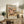 May include: Framed vintage-style beach scene poster displayed on rustic wooden sideboard in neutral-toned living room. Artwork shows people with colorful umbrellas on sandy beach in warm, muted tones. Scene includes fern plant, ceramic vase with olive branches, stacked books, and cream upholstered chair with leather throw blanket. Natural lighting creates soft shadows on white textured wall.