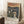 May include: Framed art print leaning against white wall on textured white rug, showing expressionist painting of anxious figures in dark coats with dramatic swirling sky in orange and red tones. Wooden side table with coffee cup visible nearby, natural window light creating soft shadows across the scene.