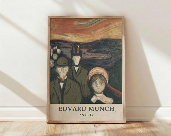 May include: Framed art print of Edvard Munch's "Anxiety" painting showing three figures in dark clothing against a swirling orange and dark blue sky. The print displays pale, haunting faces with wide eyes, set in a thin wooden frame. Positioned on light wooden flooring against a white textured wall with natural lighting creating soft shadows.