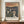May include: Framed reproduction of Munch's "Anxiety" painting showing three pale figures with haunting expressions against a swirling orange and dark blue sky. The poster has cream matting in a thin brown frame, displayed on a wooden credenza beside a white ceramic vase and stacked books against beige walls with white trim.