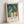 May include: Framed art print leaning against white wall showing stylized figures in teal-green tones - several nude male forms in water with flowing, elongated bodies in yellow and coral hues. The artwork features Munch's characteristic expressionist style with soft, dreamlike quality. Print has white border with artist name and title text below image, displayed in light wooden frame.