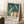 May include: Framed art print leaning against white wall on fluffy white rug, showing stylized figures in teal water - some swimming, others standing on shore in cream and coral tones. Light wood frame, cozy room setting with coffee cup on wooden surface and soft lighting from window.
