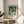 May include: Framed art print displaying swimmers in teal water with figures diving and standing on shore in yellow and coral tones, sitting on round wooden side table next to beige upholstered chair with plant shadows cast across white wall in bright natural lighting.