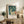 May include: Framed art print displayed on rustic wooden sideboard in neutral living room. Print shows stylized figures in teal and cream tones with text below. Scene includes fern plant, stacked books, ceramic vase, brown leather chair, and soft natural lighting creating cozy home decor atmosphere.
