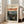 May include: A framed art print leaning against a white wall next to a beige sectional sofa in a bright living room. The poster features Edvard Munch's painting showing a figure in dark clothing against a railing with an orange-red streaked sky and water landscape in the background. The print has a cream-colored border with black text at the top, displayed in a light wood frame on hardwood flooring near large windows.