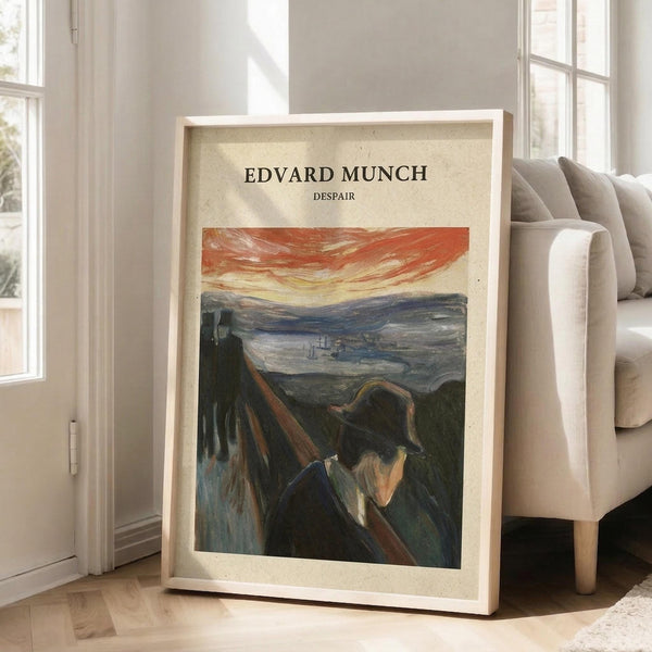 May include: A framed art print leaning against a white wall next to a beige sectional sofa in a bright living room. The poster features Edvard Munch's painting showing a figure in dark clothing against a railing with an orange-red streaked sky and water landscape in the background. The print has a cream-colored border with black text at the top, displayed in a light wood frame on hardwood flooring near large windows.