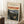 May include: Framed art print leaning against white wall corner on textured cream rug. Print shows Expressionist painting of solitary figure in dark clothing on wooden boardwalk with dramatic red-orange sky over water and landscape. Light wood frame, displayed beside wooden side table with white coffee cup and saucer, partial view of beige textured throw blanket.