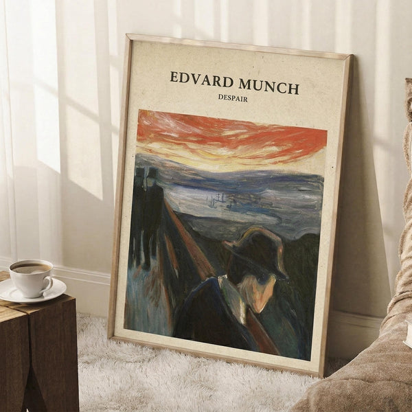 May include: Framed art print leaning against white wall corner on textured cream rug. Print shows Expressionist painting of solitary figure in dark clothing on wooden boardwalk with dramatic red-orange sky over water and landscape. Light wood frame, displayed beside wooden side table with white coffee cup and saucer, partial view of beige textured throw blanket.
