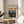 May include: Framed art print of Edvard Munch's Despair painting displayed on a round wooden side table next to a beige upholstered chair. The poster features a figure in dark clothing against a dramatic landscape with swirling orange and red sky over blue water. Natural sunlight streams across the neutral-toned room, casting soft shadows from a plant's leaves onto white walls.