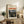 May include: Framed art print of Munch's Despair painting leaning against white wall on rustic wooden console table with drawers. Print shows figure in dark clothing against dramatic landscape with orange-red sky. Scene includes fern plant, stacked books, ceramic vase, and partial view of beige upholstered chair with brown leather pillow. Warm, natural lighting creates cozy living room atmosphere.