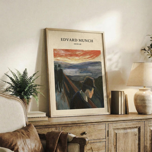 May include: Framed art print of Munch's Despair painting leaning against white wall on rustic wooden console table with drawers. Print shows figure in dark clothing against dramatic landscape with orange-red sky. Scene includes fern plant, stacked books, ceramic vase, and partial view of beige upholstered chair with brown leather pillow. Warm, natural lighting creates cozy living room atmosphere.
